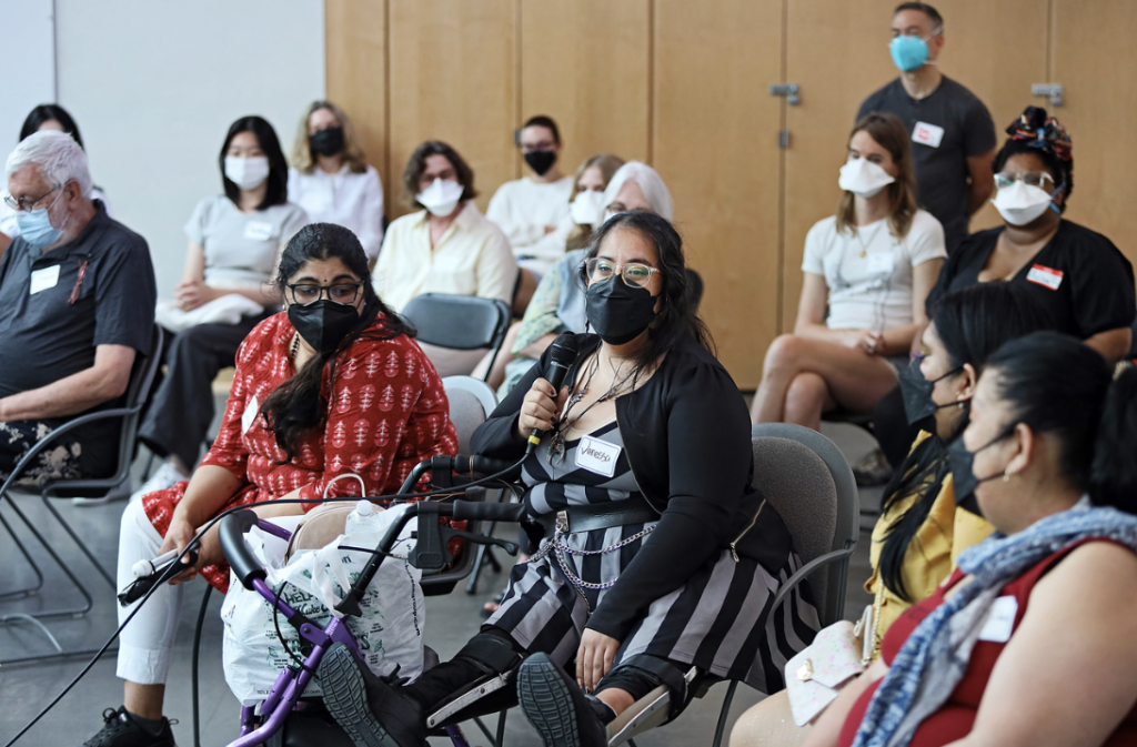 A group of individuals are seated listening to a participant introducing themselves.